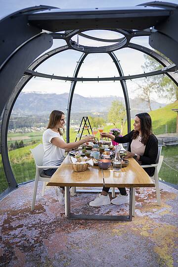 2 Frauen sitzen an einem gedecktem Tisch in der Glaskuppel und stoßen mit zwei Getränken an