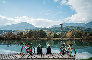 2 Personen rasten am Ufer der Drau mit ihren Gravel Bikes