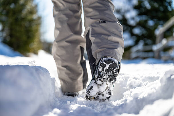 Nahaufnahme von Winterstiefeln mit Grödeln beim Gehen durch tiefen Schnee abseits der Piste.