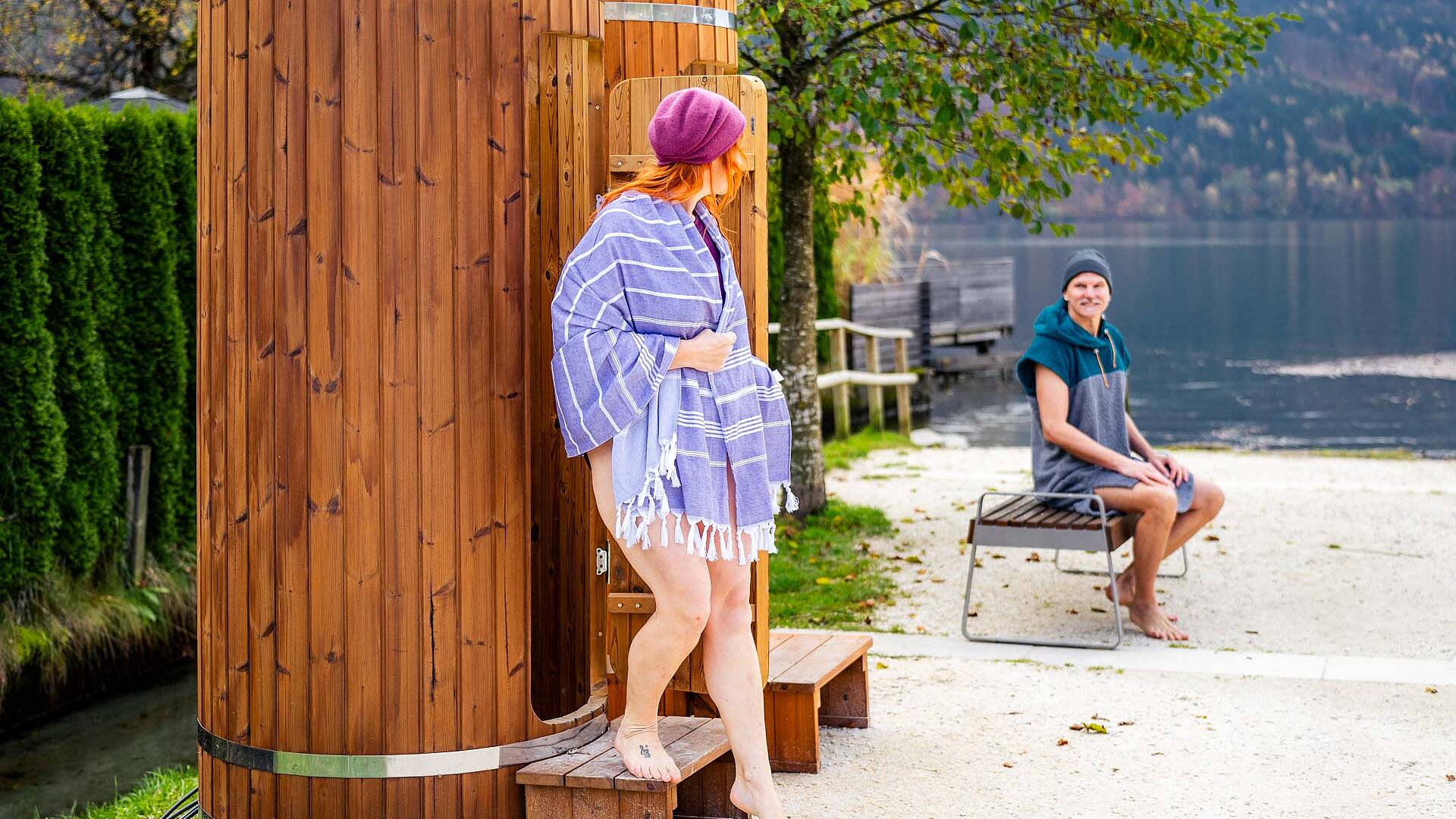 Frau steigt gerade aus Kaltbade-Kabine Millstätter See und Mann sitzt mit Badeponcho