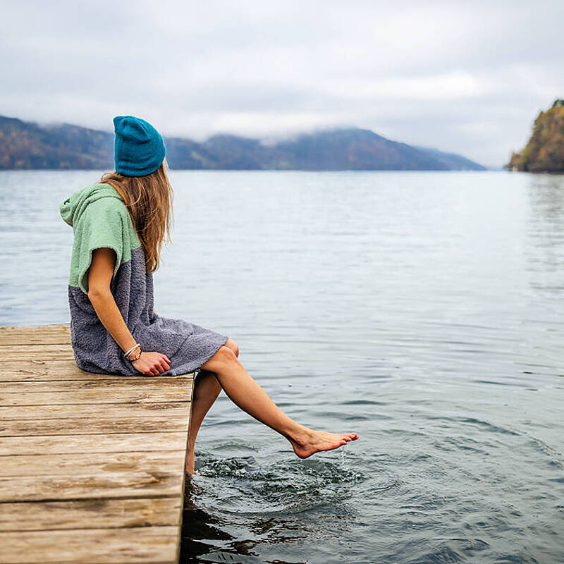 Frau sitzt mit Badeponcho auf Steg am Millstätter See und hält ihre Füße ins Wasser