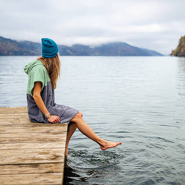 Frau sitzt mit Badeponcho auf Steg am Millstätter See und hält ihre Füße ins Wasser