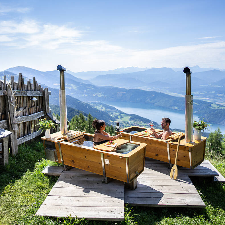 Zwei Personen genießen ein AlmBad in den Outdoorbadewannen auf der Alexanderalm, umgeben von unberührter Natur und Blick auf die Berge.