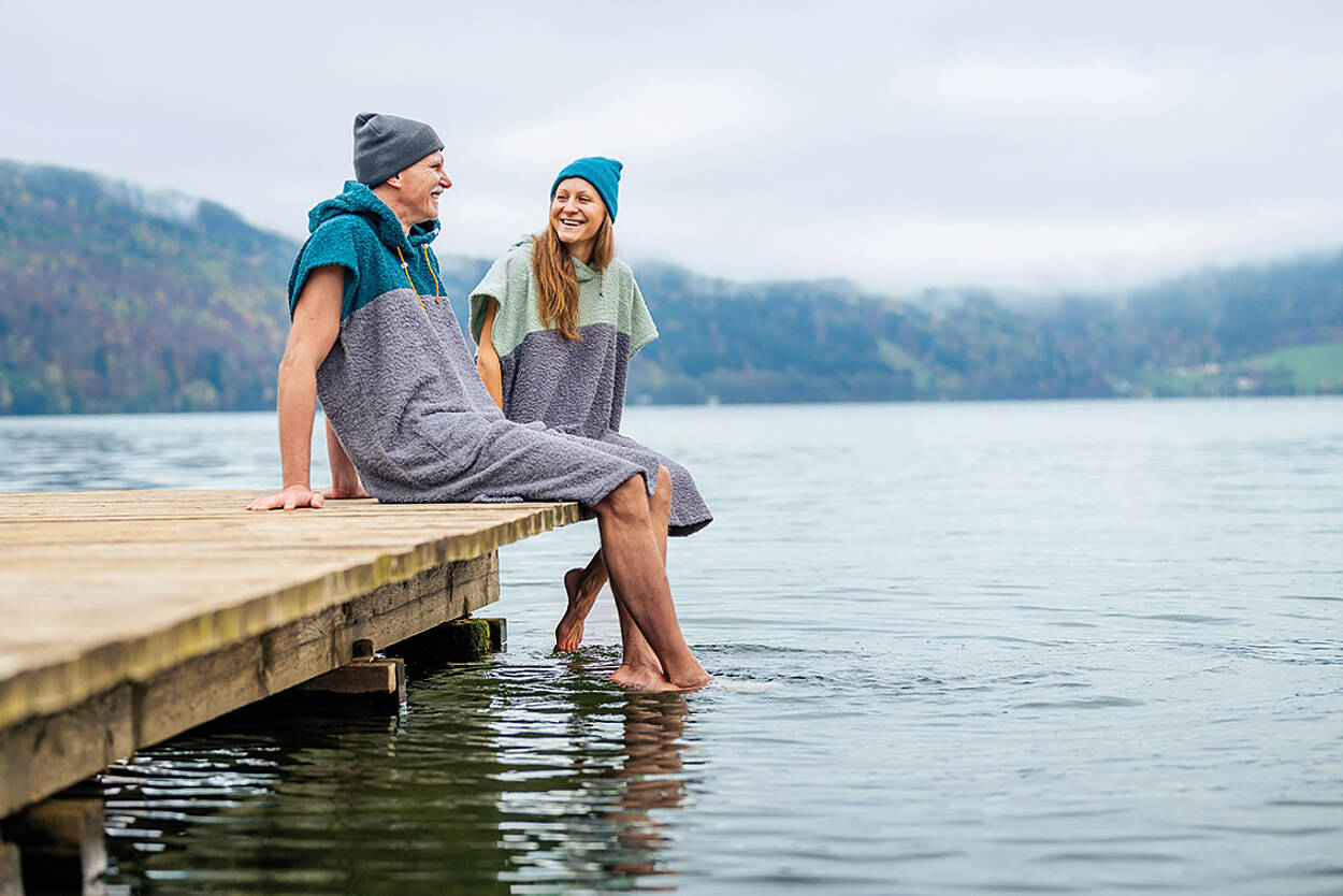 2 Personen sitzen im Badeponcho am Holzsteg am Millstätter See und halten ihre Füße in den See