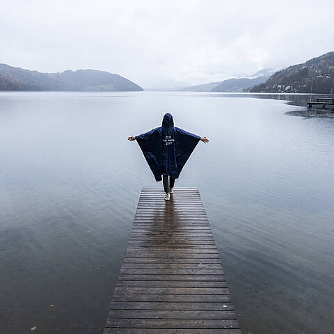 Person geht mit Regenponcho und ausgebreiteten Armen auf Steg