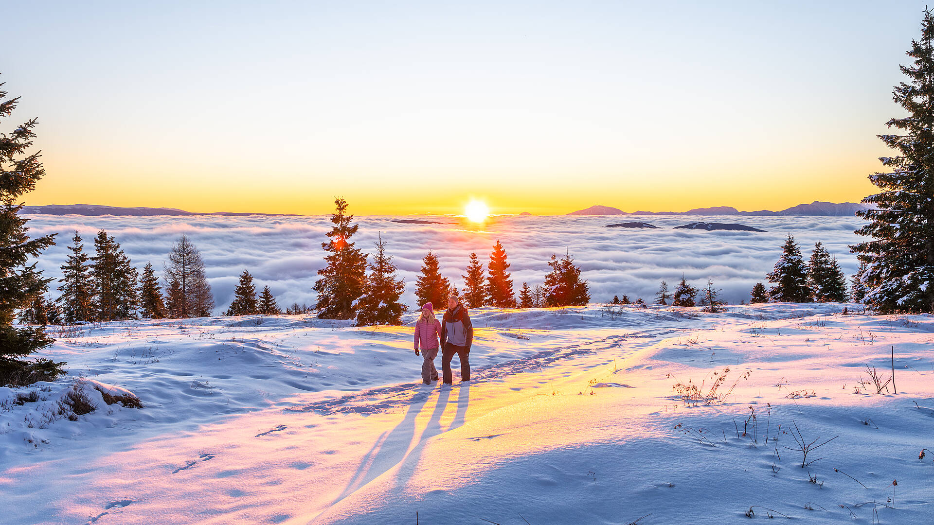 2 Personen stehen im warmen Abendlicht auf verschneitem Plateau mit Blick über die winterlichen Nockberge.